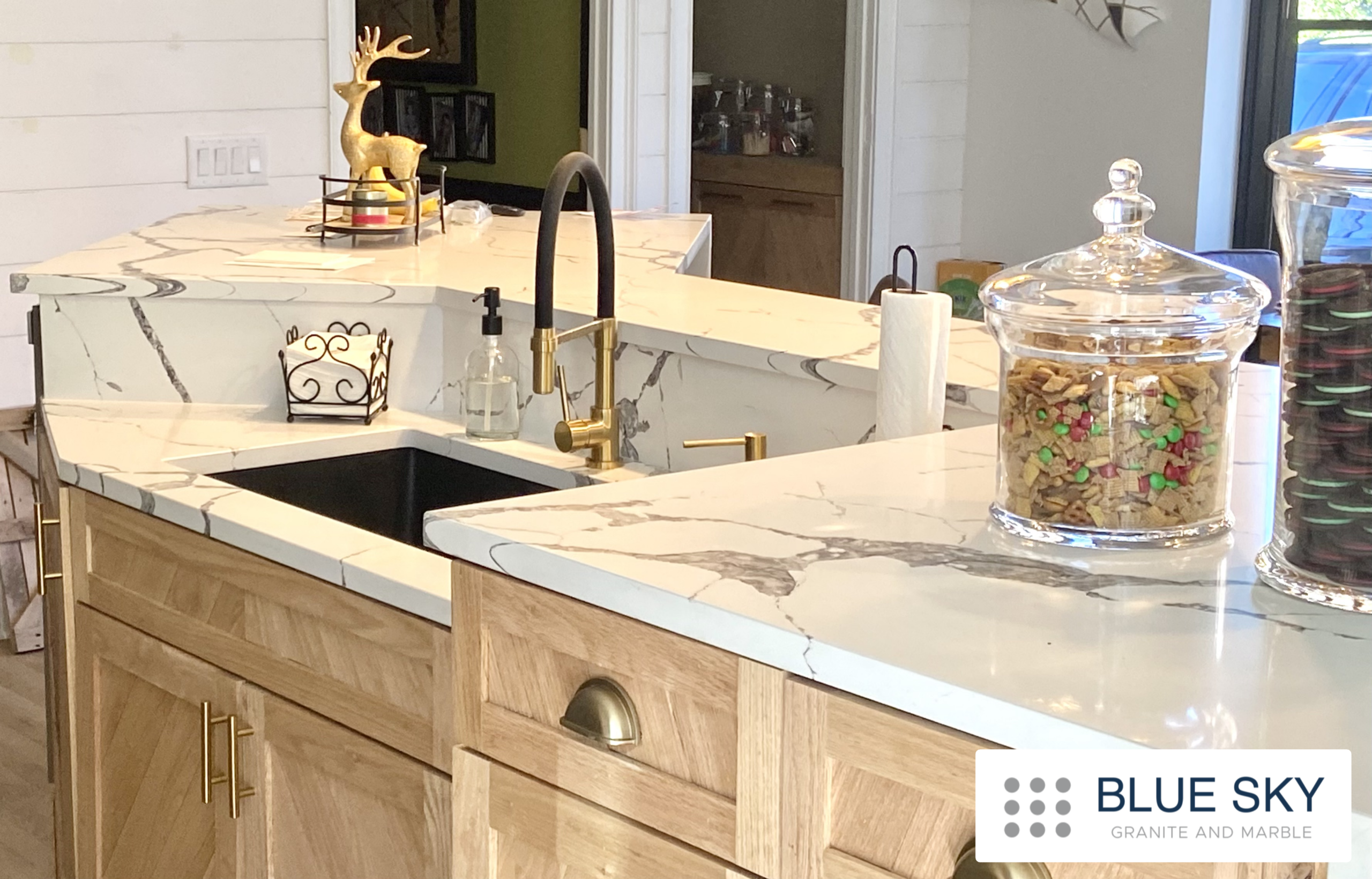 White veined quartz island with a sink and black-and-brass faucet in a kitchen, with the Blue Sky Granite and Marble logo inside the lower right.